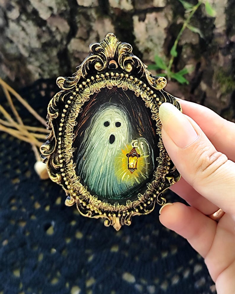 May include: A miniature painting of a ghost with a lantern, set within an ornate gold and black frame. The ghost is depicted with a surprised expression. The frame is held by a hand, showcasing the intricate details of the artwork.