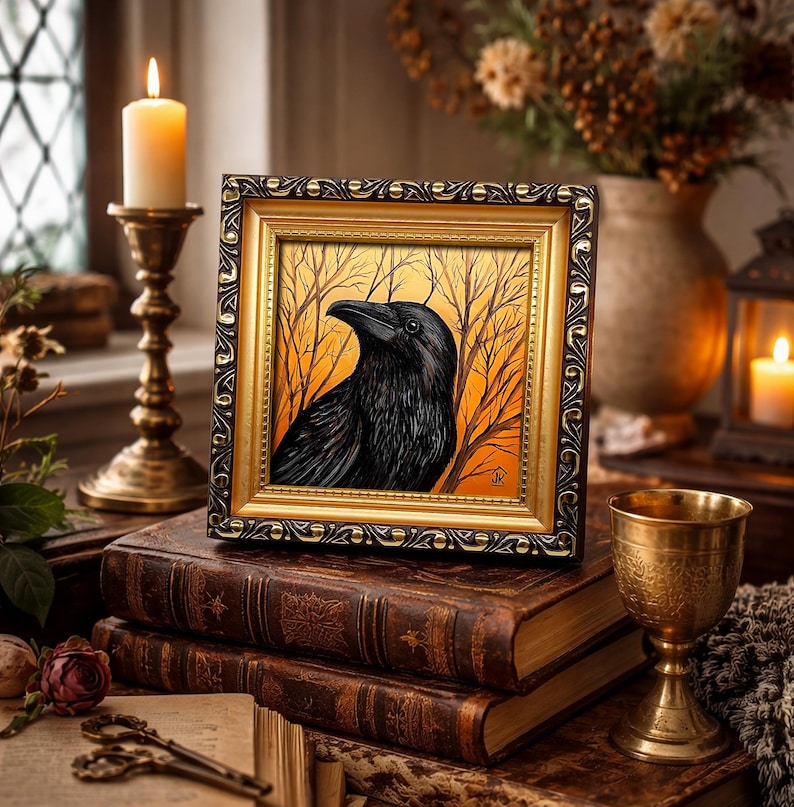 Pode incluir: Uma obra de arte emoldurada com um corvo preto contra um fundo de &aacute;rvores nuas e um c&eacute;u laranja. A moldura &eacute; dourada com uma borda marrom escura. A obra de arte repousa sobre uma pilha de livros antigos.