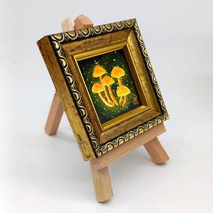 May include: A small framed painting of glowing orange mushrooms on a dark green background. The painting is on a wooden easel.