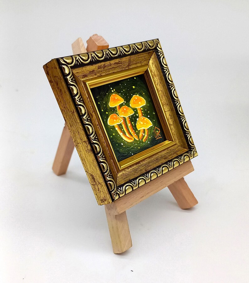 May include: A small framed painting of glowing orange mushrooms on a dark green background. The painting is on a wooden easel.