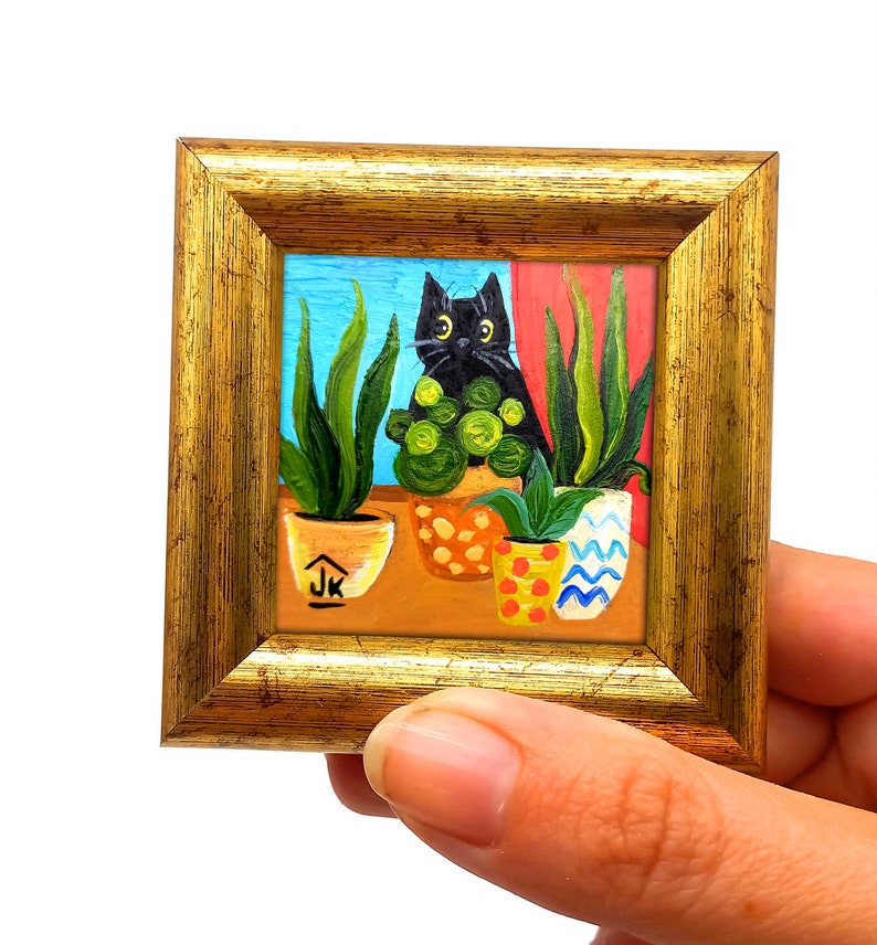 May include: A framed painting of a black cat sitting in front of a window with potted plants. The plants are in colorful pots with a blue, white, and red pattern. The cat is looking at the viewer with green eyes. The painting is signed "JK" in the bottom right corner.