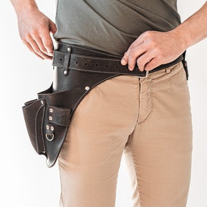 May include: A dark brown leather tool belt with multiple pockets and a belt loop. The belt has white stitching and a silver buckle. The tool belt is worn over tan pants and a gray shirt.