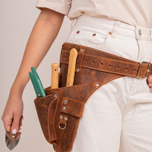 May include: A brown leather gardening tool belt with multiple pockets and a belt buckle. The belt holds a pair of green handled shears, two wooden handled tools, and a silver trowel. The belt is worn around the waist.