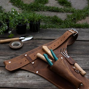 May include: A brown leather gardening tool belt featuring multiple pockets and loops, secured with a metal buckle. The belt holds a small hand rake and two tools with teal handles, constructed with visible stitching and silver-toned metal rivets.