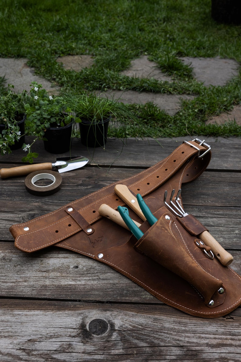 May include: A brown leather gardening tool belt featuring multiple pockets and loops, secured with a metal buckle. The belt holds a small hand rake and two tools with teal handles, constructed with visible stitching and silver-toned metal rivets.
