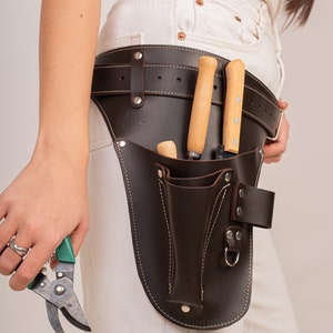 May include: A dark brown leather tool belt featuring a wide waist strap with metal rivets, a holster for two wooden-handled tools, a smaller front pocket, and a metal D-ring. The belt is worn around the waist and includes visible stitching.