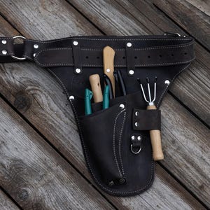 May include: A dark brown leather tool belt featuring multiple pockets and silver-toned metal rivets. The belt contains several gardening tools with wooden handles, including a small rake and knives, secured within the integrated leather pouches.