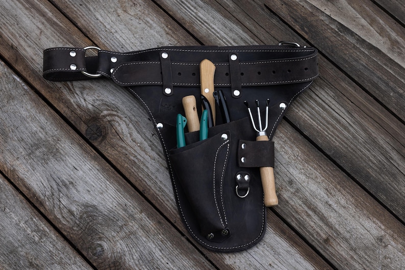 May include: A dark brown leather tool belt featuring multiple pockets and silver-toned metal rivets. The belt contains several gardening tools with wooden handles, including a small rake and knives, secured within the integrated leather pouches.