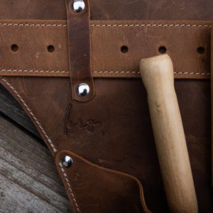 May include: A brown leather tool belt featuring silver-toned metal rivets and white stitching. Two wooden-handled tools are tucked into the pockets. A small, embossed signature is visible on the leather surface near the center of the belt.