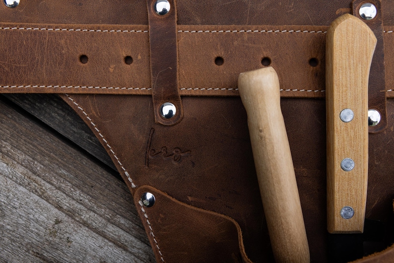 May include: A brown leather tool belt featuring silver-toned metal rivets and white stitching. Two wooden-handled tools are tucked into the pockets. A small, embossed signature is visible on the leather surface near the center of the belt.