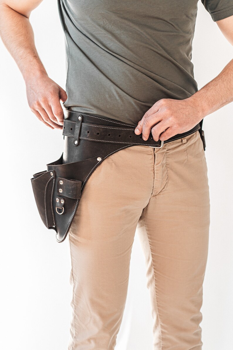 May include: A dark brown leather tool belt with multiple pockets and a belt loop. The belt has white stitching and a silver buckle. The tool belt is worn over tan pants and a gray shirt.