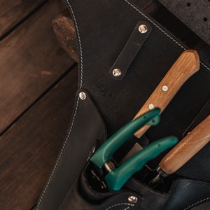 May include: Black leather gardening tool belt with white stitching, holding green handled pruning shears and two wooden handled tools. The belt has silver rivets and a metal ring.