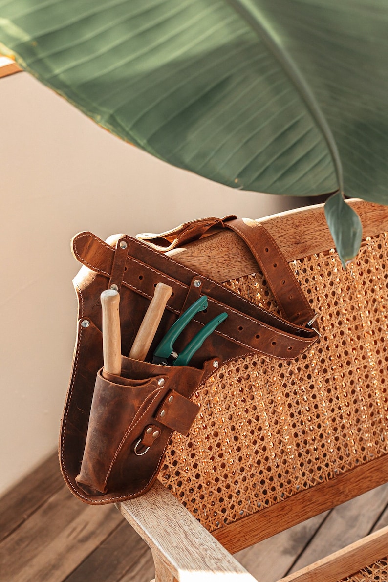 May include: A brown leather tool belt featuring multiple pockets and an adjustable strap. The belt contains two wooden-handled tools and a pair of green-handled pruning shears. The leather is secured with metal rivets and includes a small metal ring.