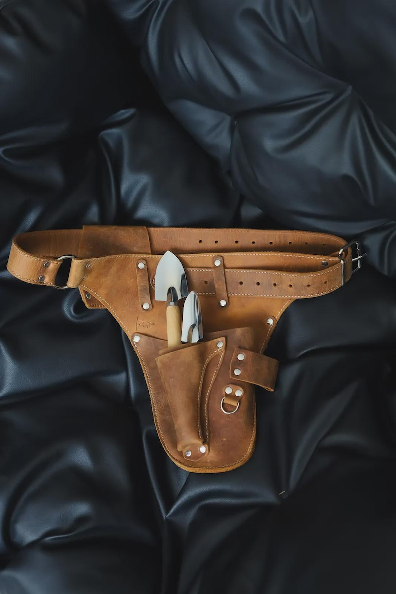 May include: A tan leather tool belt featuring an adjustable strap with metal buckle holes, silver-toned rivets, and a holster pocket containing two small gardening hand tools with wooden handles and polished metal heads, secured by a metal ring.