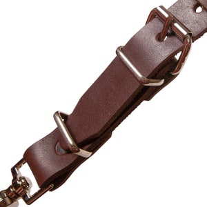 May include: Close-up of a brown leather strap with silver metal buckles. The strap is folded over itself and the buckles are visible.