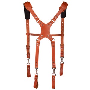 May include: A brown leather camera strap with adjustable straps and metal buckles. The strap has a Y-shaped design and is designed to be worn over the shoulders.