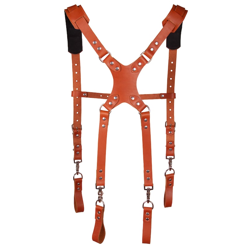 May include: A brown leather camera strap with adjustable straps and metal buckles. The strap has a Y-shaped design and is designed to be worn over the shoulders.