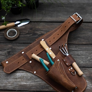 May include: A brown leather gardening tool belt featuring an adjustable strap with a metal buckle, multiple pockets, and silver-tone rivets. The belt holds a small hand rake with a wooden handle and two small hand tools with teal handles.