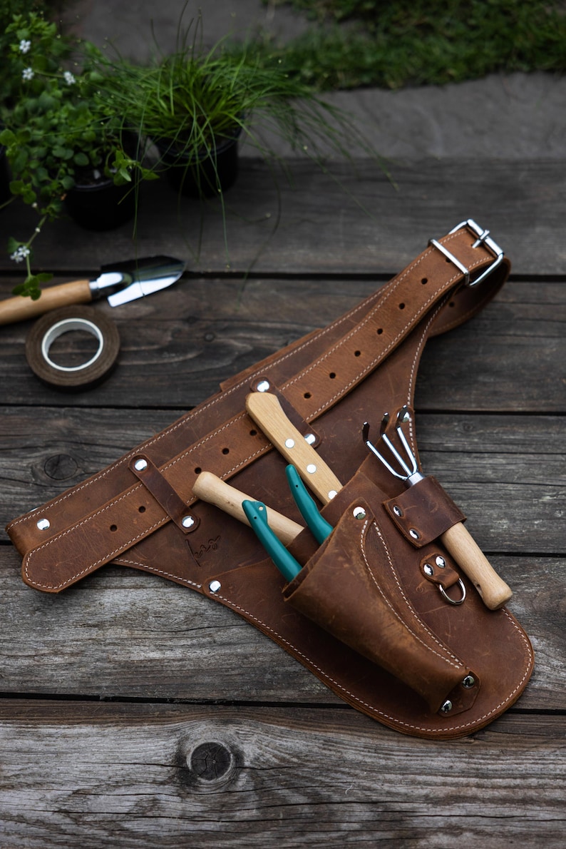 May include: A brown leather gardening tool belt featuring an adjustable strap with a metal buckle, multiple pockets, and silver-tone rivets. The belt holds a small hand rake with a wooden handle and two small hand tools with teal handles.