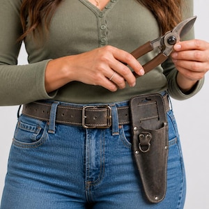 Op de afbeelding: Een snoeischaar met bruine handgrepen en een leren holster bevestigd aan een bruine riem. De riem heeft een grote vierkante gesp. De persoon draagt een blauwe spijkerbroek en een groen shirt met lange mouwen.