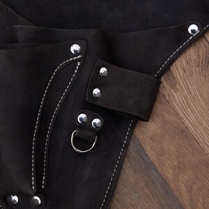 May include: A black leather tool holster featuring multiple pockets, silver-toned metal rivets, and a sturdy D-ring attachment. The accessory is constructed with reinforced stitching along the edges and includes several loops for secure storage.