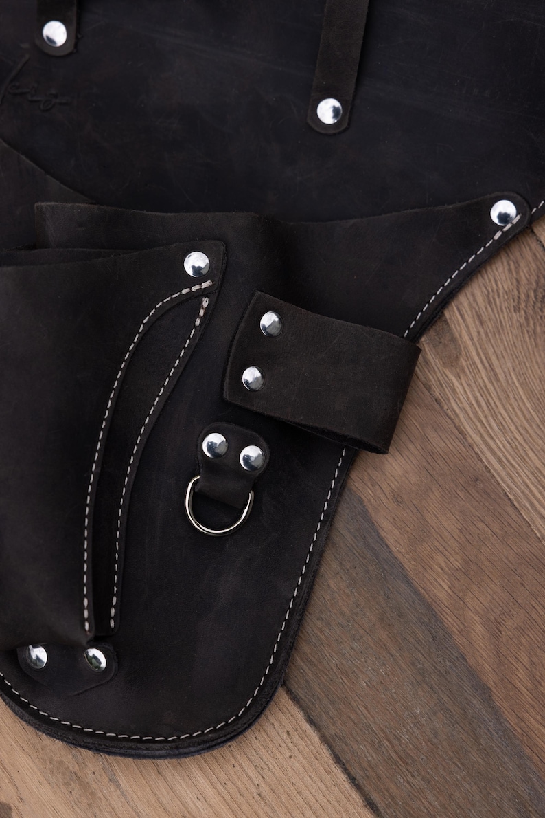 May include: A black leather tool holster featuring multiple pockets, silver-toned metal rivets, and a sturdy D-ring attachment. The accessory is constructed with reinforced stitching along the edges and includes several loops for secure storage.
