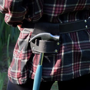 May include: A black leather tool belt worn around the waist over a red, black, and white plaid flannel shirt. The belt features a dedicated holster holding a metal hammer with a blue textured handle, secured with silver-toned metal rivets.