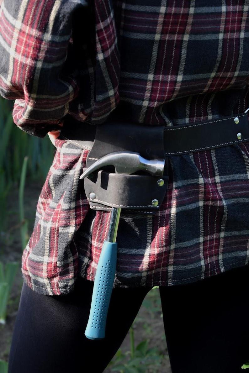 May include: A black leather tool belt worn around the waist over a red, black, and white plaid flannel shirt. The belt features a dedicated holster holding a metal hammer with a blue textured handle, secured with silver-toned metal rivets.