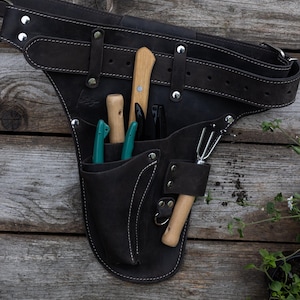 May include: A dark brown leather tool belt featuring a wide adjustable strap with silver metal rivets and multiple pockets. The belt holds several gardening tools with wooden and teal handles, including a small hand rake and various pruning blades.