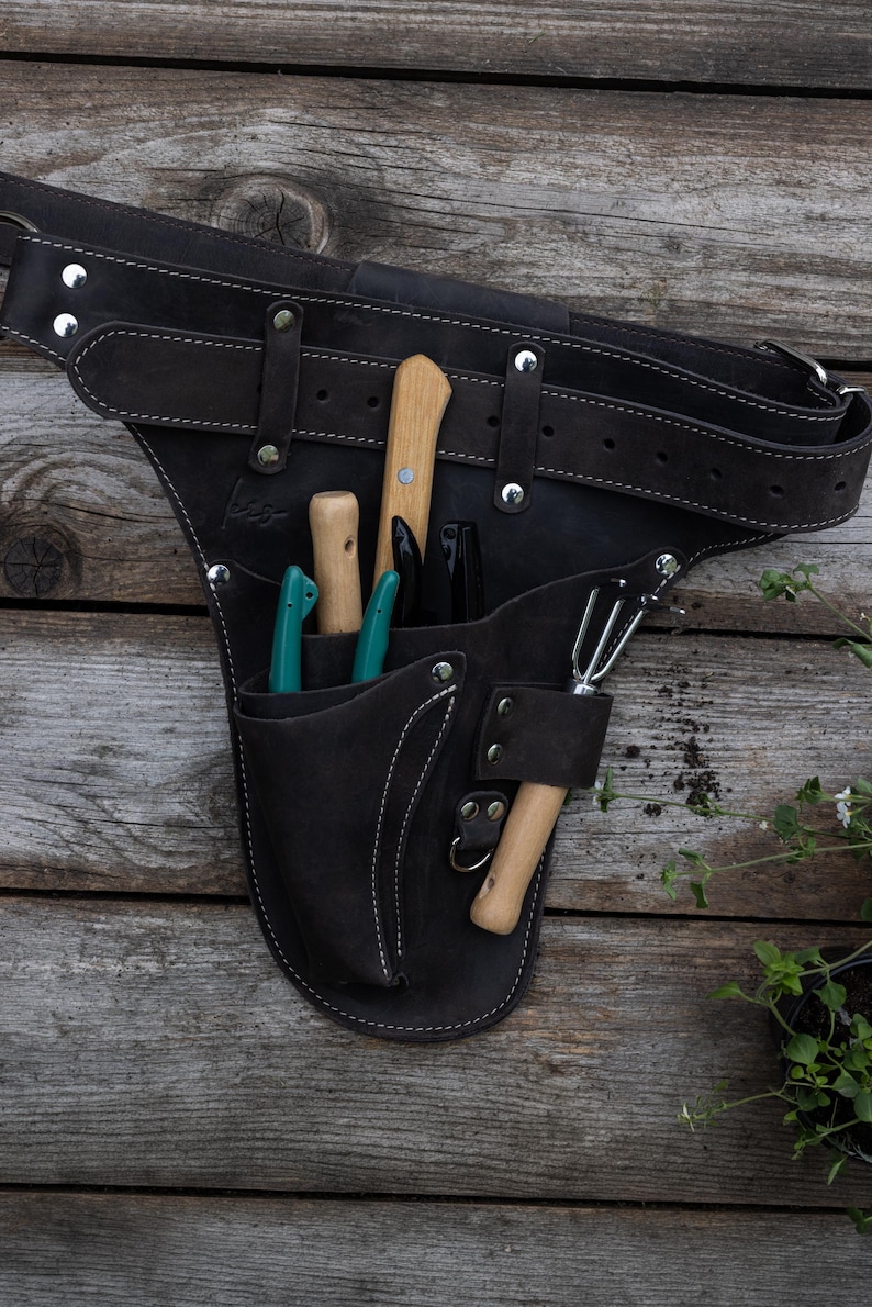 May include: A dark brown leather tool belt featuring a wide adjustable strap with silver metal rivets and multiple pockets. The belt holds several gardening tools with wooden and teal handles, including a small hand rake and various pruning blades.