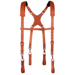 May include: A brown leather camera strap with adjustable straps and metal buckles. The strap is designed to be worn over the shoulders and has a chest strap for added support.