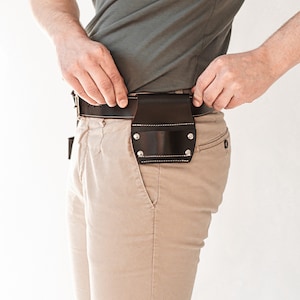 May include: A dark brown leather belt holster attached to a matching dark brown leather belt. The holster features a rectangular pouch design with visible silver-toned metal rivets at the corners and light-colored stitching along the edges.