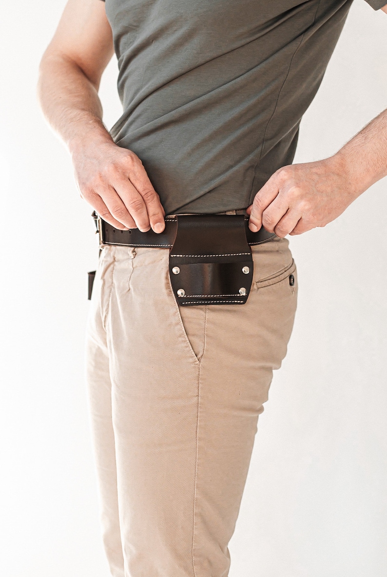 May include: A dark brown leather belt holster attached to a matching dark brown leather belt. The holster features a rectangular pouch design with visible silver-toned metal rivets at the corners and light-colored stitching along the edges.