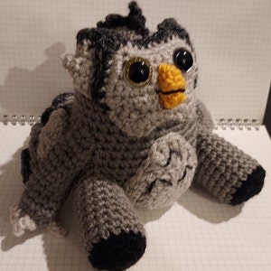 May include: A gray crocheted owl plush toy with black accents and large, round, glittery eyes. The owl has a yellow beak and is sitting on a white surface.