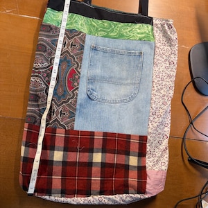 May include: A patchwork tote bag with black handles. The bag is made from various fabrics, including denim, paisley, plaid, and floral prints. A measuring tape shows the bag is approximately 29 inches long. The bag has a denim pocket.