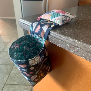 May include: A fabric sewing organizer with a hanging design. The exterior features a navy blue background with sewing-themed patterns, while the interior and a small pillow have floral patterns. The organizer is designed to hang over a countertop.