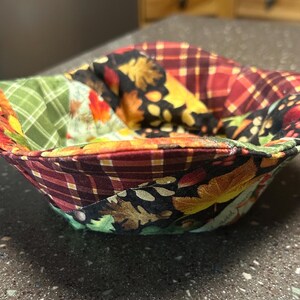 Może przedstawiać: Ręcznie robiony przytulny kubek z patchworkowym wzorem. Miska ma różne jesienne tkaniny, w tym kratę, liście i kwiatowe wzory w odcieniach czerwieni, pomarańczy, żółci i zieleni. Przytulny kubek jest przeznaczony do trzymania miski z gorącym jedzeniem.