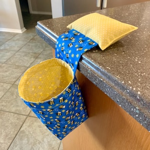May include: A blue fabric trash can with a yellow bee pattern hangs from a kitchen counter. The can has a yellow fabric lining. A yellow pillow with white polka dots is tucked into the top of the can.