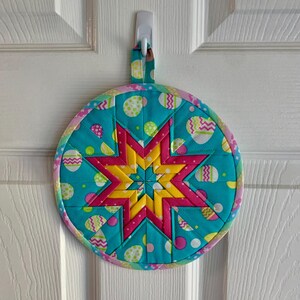 May include: A round, quilted hot pad with a starburst design in pink, yellow, and blue. The fabric is patterned with pastel Easter eggs and polka dots.