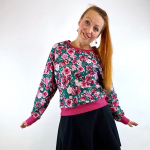 Könnte beinhalten: Eine Frau trägt einen grünen und pinken floralen Sweatshirt mit einem schwarzen Rock und schwarzen kniehohen Stiefeln.