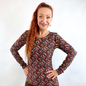 Könnte beinhalten: Ein langärmliges Oberteil mit dunkelbraunem Hintergrund und einem bunten Blumenmuster. Das Oberteil wird mit einer braunen Hose kombiniert. Die Frau lächelt und hat lange rote Haare.