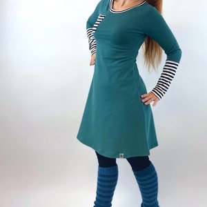 Puede incluir: Un vestido de manga larga azul turquesa con un detalle de rayas blancas y negras en las mangas y un peque&ntilde;o bolsillo en el pecho. El vestido se lleva con polainas de rayas azules y zapatillas negras.