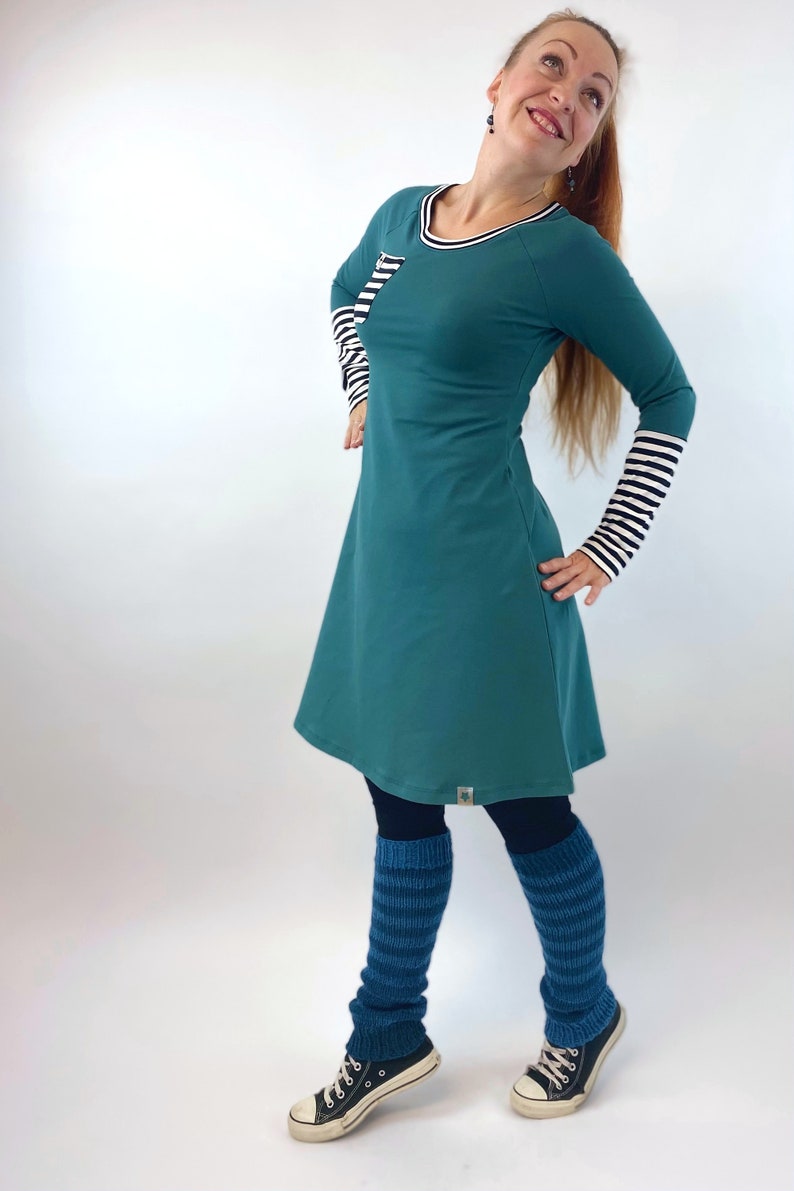 Puede incluir: Un vestido de manga larga azul turquesa con un detalle de rayas blancas y negras en las mangas y un peque&ntilde;o bolsillo en el pecho. El vestido se lleva con polainas de rayas azules y zapatillas negras.