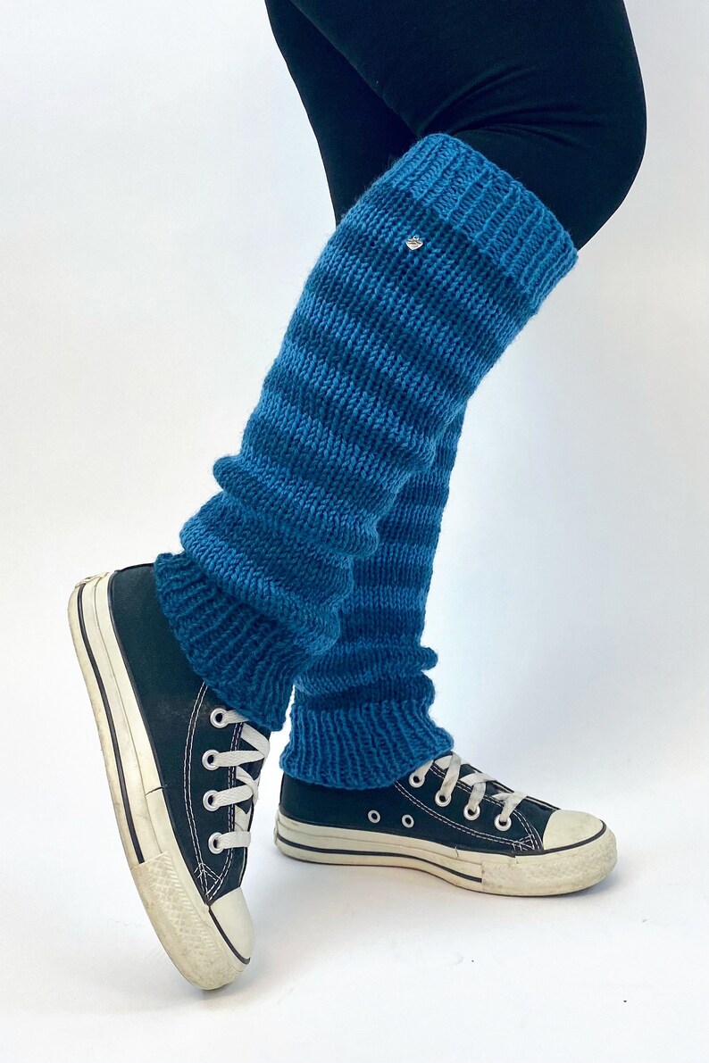Puede incluir: Calentadores de piernas azules a rayas hechos de tejido de punto. Los calentadores de piernas se llevan sobre leggings negros y zapatillas negras.