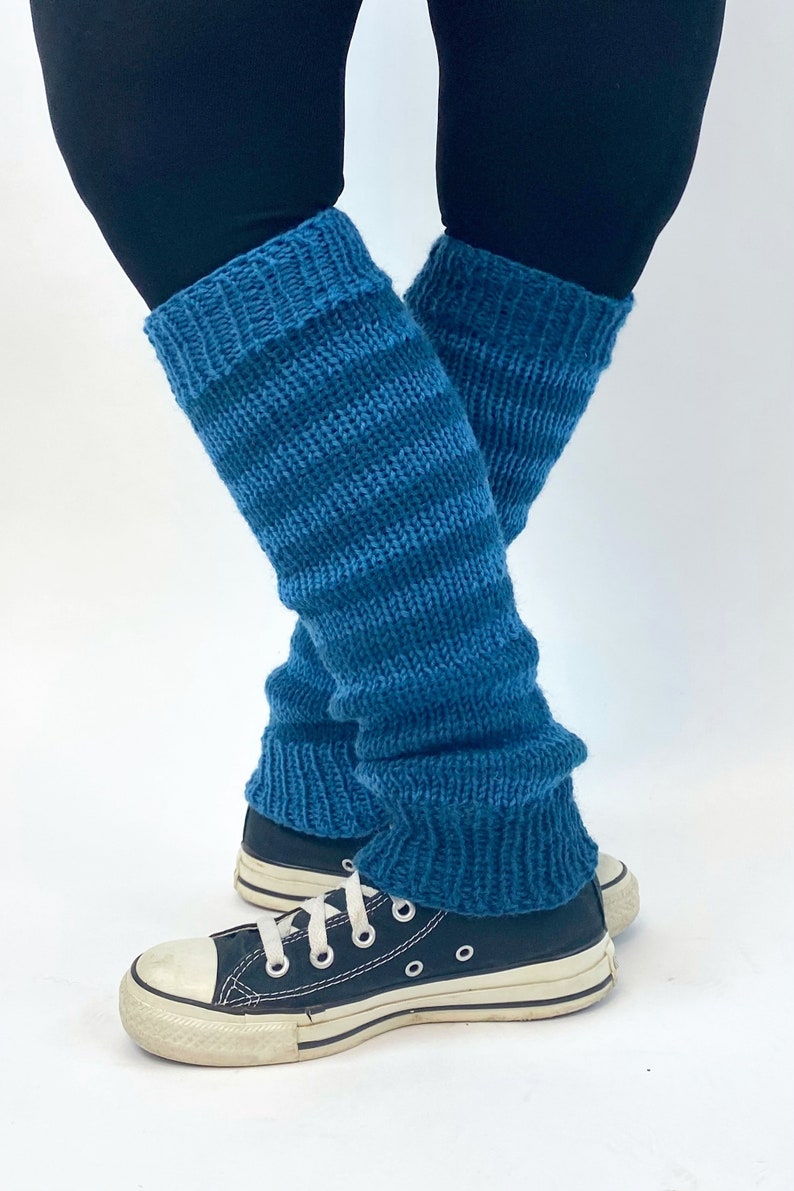 Puede incluir: Un par de polainas de punto azul turquesa con rayas blancas. Las polainas se llevan sobre unos leggings negros y unas zapatillas negras.