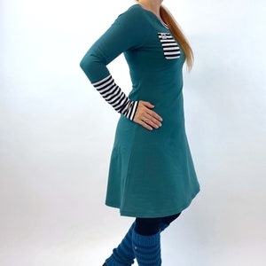 Puede incluir: Un vestido de manga larga azul turquesa con un bolsillo a rayas y pu&ntilde;os a rayas blanco y negro. El vestido tiene una falda acampanada y se lleva con polainas azules y zapatillas negras.