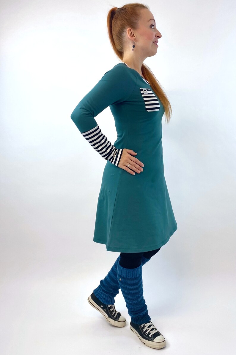 Puede incluir: Un vestido de manga larga azul turquesa con un bolsillo a rayas y pu&ntilde;os a rayas blanco y negro. El vestido tiene una falda acampanada y se lleva con polainas azules y zapatillas negras.