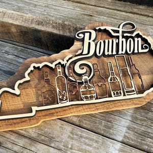 May include: Wooden sign shaped like the state of Kentucky with the word "Bourbon" in a stylized font. The sign features a silhouette of multiple bottles of bourbon.