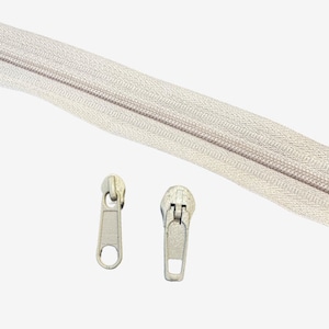 May include: A close-up of a beige zipper with two silver zipper pulls. The zipper is lying flat on a white surface.