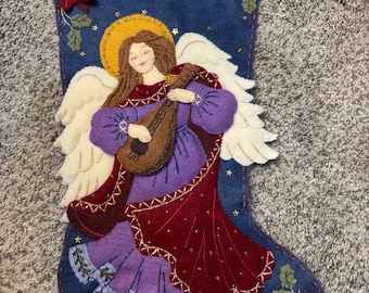 Puede incluir: Un calcetín navideño hecho a mano de fieltro azul marino presenta un ángel tocando un laúd. El ángel viste una túnica morada y burdeos con alas blancas y un halo dorado. El calcetín está decorado con acebo, estrellas y flores de pascua rojas.